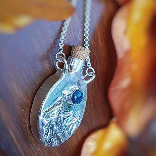 Silver pendant bottle with blue gemstone on a wooden surface, fantasy inspired