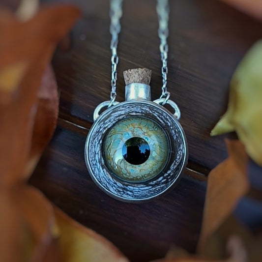 Necklace with a detailed green glass eye-shaped corked jar pendant on a wooden surface. Perfect for Spooky season, Halloween, Autumn, all the 'ber' months.